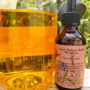 May include: A brown glass bottle with a black dropper cap and a label that reads "Anne's Backyard Herbal Oil Infusion." The bottle is next to a large glass jar filled with a golden-colored liquid. The label lists ingredients and usage instructions.