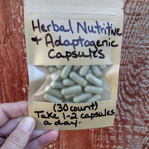Pu&ograve; includere: Un piccolo sacchetto di carta marrone con una finestra di plastica trasparente che contiene una quantit&agrave; di capsule verde chiaro. Il sacchetto ha un testo scritto a mano che dice "Herbal Nutritive + Adaptogenic Capsules (30 count) Take 1-2 capsules a day."