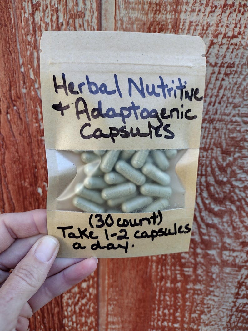 Pu&ograve; includere: Un piccolo sacchetto di carta marrone con una finestra di plastica trasparente che contiene una quantit&agrave; di capsule verde chiaro. Il sacchetto ha un testo scritto a mano che dice "Herbal Nutritive + Adaptogenic Capsules (30 count) Take 1-2 capsules a day."