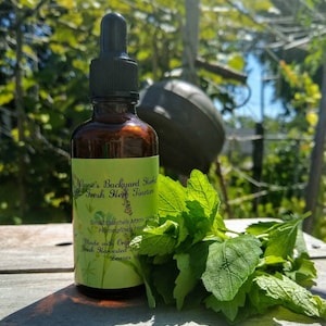 May include: A brown glass bottle with a dropper top and a label that reads "Anne's Backyard Herbal Fresh Herb Tincture". The label also includes the text "Small Batches Made from Homegrown Herbs", "Made with Organic", "Fresh Harvested", and "Leaves of". A bunch of green leaves are in the foreground.