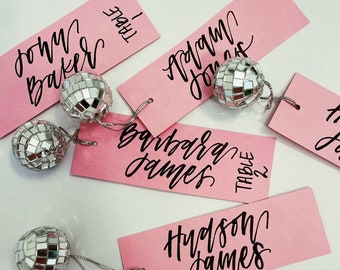 May include: Pink table cards with black calligraphy names and the words "Table 1" and "Table 2". Each card has a silver disco ball attached to it.