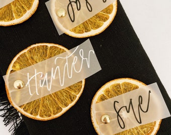 May include: Four dried orange slices with gold accents and white paper place cards with black calligraphy names:  'Josh', 'Hunter', 'Eve', and 'Jim'.