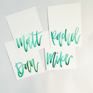 Puede incluir: Cuatro tarjetas blancas con nombres escritos en acuarela verde. Los nombres son "Matt", "Rachel", "Dan" y "Mike".