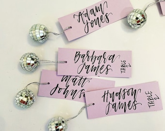 May include: Pink place cards with black calligraphy names and the words "Table 1" and "Table 2" on them. Each place card has a small silver disco ball attached to it with a silver string.