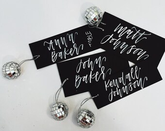 May include: Four black place cards with white calligraphy names, "Anna Baker", "John Baker", "Matt Johnson", and "Kendall Johnson". Each card has a silver disco ball attached to it with a string.