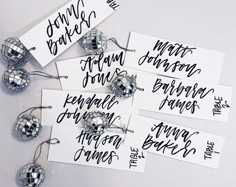 May include: White place cards with black calligraphy names and table numbers. The names are John Baker, Adam Jones, Matt Johnson, Barbara James, Kendall Johnson, Hudson James, and Anna Baker. The table numbers are 1, 2, and 3. There are silver disco ball ornaments attached to each place card.