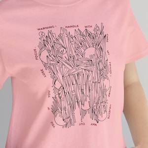 May include: Pink t-shirt with a black and white graphic design. The graphic features a variety of geometric shapes and the text "WARNING! HANDLE WITH CARE. MAY CUT YOU. IF YOU LOSE FOCUS, MY PRINTS MAY ALWAYS OBJECT."