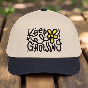 May include: A beige baseball cap with a navy blue peak. The cap displays the words "Keep Growing" in black script, accompanied by a yellow flower graphic. The cap is made of a durable fabric and has a curved peak.