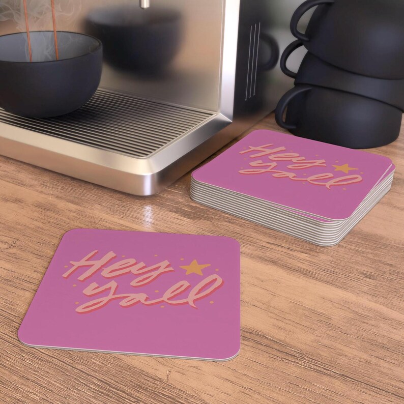 May include: Two pink square coasters with the text "Hey y'all" in a pink and orange font with a gold star.