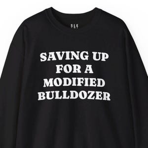 May include: Black sweatshirt with the text "SAVING UP FOR A MODIFIED BULLDOZER" in white block letters. The sweatshirt has a crew neck and long sleeves. The brand logo "SLE" is at the top.