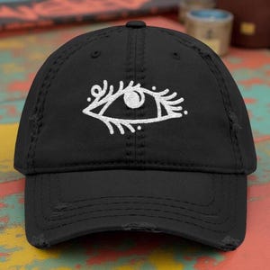 May include: Black distressed baseball cap featuring a white embroidered eye design on the front. The cap has a curved brim and a worn, vintage look. The eye design is detailed with eyelashes and decorative elements.