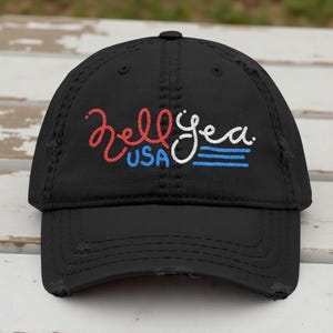 Könnte beinhalten: Schwarze Baseballkappe mit Used-Look und dem gestickten Text "Kelly Yea USA" in Rot, Weiß und Blau. Die Kappe hat einen gebogenen Schirm und einen abgenutzten Look mit ausgefransten Kanten.