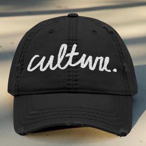 Op de afbeelding: Zwarte baseballcap met het woord "culture" in witte letters geborduurd. De cap heeft een distressed look met gerafelde randen. Het is een klassiek zes-panel ontwerp met een gebogen klep.