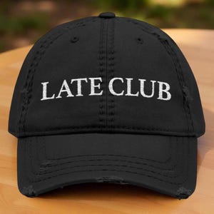May include: Black baseball cap with the embroidered text "LATE CLUB" in white. The cap has a distressed, worn look with frayed edges on the brim and crown. The cap is sitting on a wooden surface.