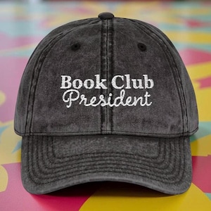May include: A dark gray baseball cap with the embroidered text "Book Club President" in white. The cap has a curved brim and a button on top. The hat is made of a soft, worn material.