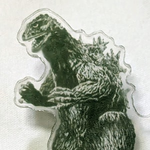 May include: A green plastic pin featuring a silhouette of Godzilla, a famous Japanese monster.