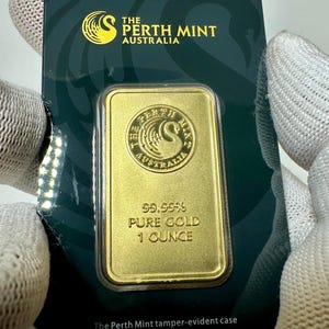 Puede incluir: Una barra de oro con el logotipo de la Perth Mint y el texto "99,99% ORO PURO 1 ONZA" sobre un fondo verde. La barra está en una caja a prueba de manipulaciones.