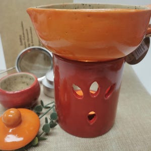 May include: An orange ceramic essential oil diffuser with a matching lid and small red ceramic bowl. The diffuser has a red base with leaf-shaped cutouts and an orange top. A metal tea strainer and tea lights are also visible.