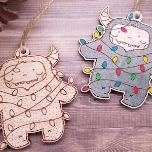 May include: Two wooden Christmas ornaments shaped like monsters wearing string lights. One monster is light brown with yellow lights and the other is gray with multi-colored lights.