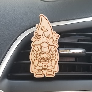 May include: A wooden gnome air freshener with a floral design and a frog on the hat. The gnome is holding a pot of flowers. The air freshener is hanging from a car's air vent.