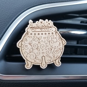May include: A wooden car air freshener shaped like a cauldron with a decorative design. The cauldron has a crown on top and is filled with swirling designs.