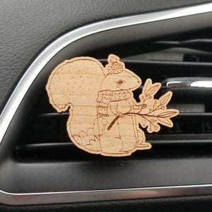 May include: A wooden car air freshener featuring a squirrel wearing a scarf and holding a sprig of holly. The squirrel is depicted in a cartoon style with a whimsical expression.