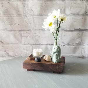 Op de afbeelding: Een kleine houten tray met een glazen vaas met witte bloemen, een turquoise konijnenbeeldje, een brandende kaars en decoratieve stenen. De tray staat op een grijs oppervlak met een witte bakstenen achtergrond.