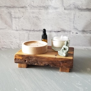 Op de afbeelding: Een houten platform met een natuurlijke rand houdt een kleine kaars in een glazen houder, een klein Boeddha-beeld, een houten wierookhouder en een klein flesje vast. De items zijn gerangschikt op een grijs oppervlak tegen een witte bakstenen muur.