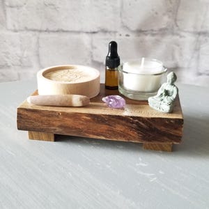Op de afbeelding: Een houten dienblad met een kleine kaars, een klein Boeddha-beeld, een kleine fles etherische olie, een klein amethistkristal en een houten kom met een stamper. Het dienblad is gemaakt van donker hout.