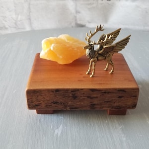 Op de afbeelding: Een kleine, vierkante houten displaystandaard met een gouden gevleugelde hertenfiguur en een gele kristal. De standaard is gemaakt van roodbruin hout en heeft een natuurlijk, getextureerd uiterlijk. Het beeldje is gedetailleerd en staat op de standaard.