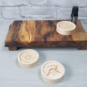 May include: A wooden tray with three round wooden coasters. The coasters feature different designs, including a sun, a moon, and a deer. A small bottle of essential oil is on the tray.