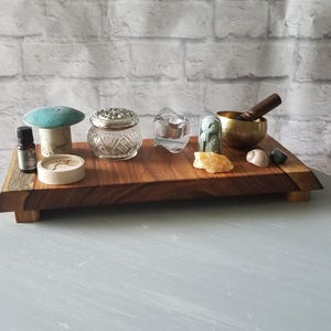 Op de afbeelding: Een houten dienblad met diverse items, waaronder een turquoise paddenstoel, een klein glazen potje, een kaars, een zangkom, kristallen en een klein flesje. Het dienblad staat op een lichtblauwe ondergrond tegen een bakstenen muur.