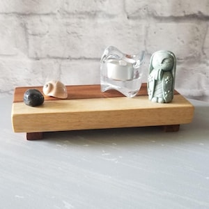 Puede incluir: Una bandeja decorativa de madera con una base de madera clara y un acento de madera oscura. Contiene una piedra negra, una pequeña concha beige, un portavelas de cristal transparente con una vela encendida y una pequeña estatua verde. La bandeja está sobre una superficie gris.
