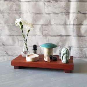 May include: A wooden tray with various decorative items. Includes a small vase with white flowers, a small bottle, a wooden coaster, a mushroom-shaped object, a small stone sculpture, and a glass candle holder. The tray is on a gray surface.