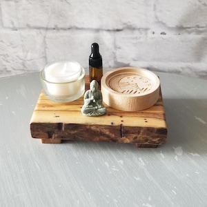 Op de afbeelding: Een houten blok met een kleine kaars in een glazen houder, een klein Boeddha-beeld, een houten onderzetter en een bruine etherische olie fles. De items zijn gerangschikt op een grijze ondergrond.