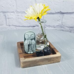 May include: A small, light blue figurine of a person with their hands clasped in prayer is sitting on a wooden tray with black stones and a small vase with a white flower.