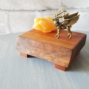 Op de afbeelding: Een kleine houten displaystandaard met een gouden gevleugelde hertenfiguur en een gele kristal. De standaard is rechthoekig met een donkere houten afwerking en staat op vier kleine pootjes. Het hertenfiguur is gedetailleerd.