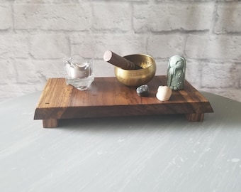 May include: A wooden meditation tray with a brass singing bowl, a wooden mallet, a small glass candle holder, a green stone owl, and two small stones. The tray is on a light gray surface.