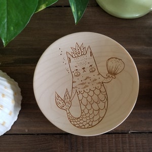 Può includere: Un piatto di legno con un'illustrazione disegnata a mano di una sirena gatto con una corona e che tiene una conchiglia. La sirena gatto ha una coda a strisce e una corona sulla testa.
