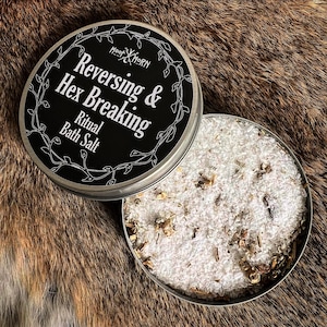 May include: A round metal tin containing white bath salt mixed with dried herbs. The lid is black with white text that reads Hoof & Horn Reversing & Hex Breaking Ritual Bath Salt, surrounded by a thin white vine illustration on the border.