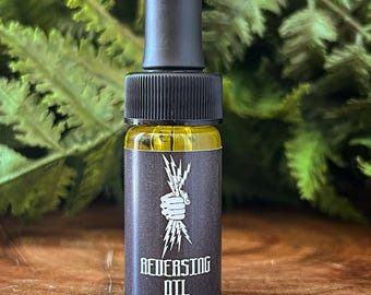 Può includere: Una piccola bottiglia di vetro con liquido giallo e tappo contagocce nero. L'etichetta è marrone scuro con una grafica bianca di una mano che tiene dei fulmini e le parole "REVERSING OIL". Lo sfondo è un fogliame verde sfocato.