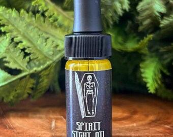 Peut inclure: Petite bouteille en verre avec un compte-gouttes noir, remplie de liquide jaune. L'étiquette affiche un squelette blanc dans un cercueil et le texte "SPIRIT SIGHT OIL". En arrière-plan, des feuilles vertes et une surface en bois.