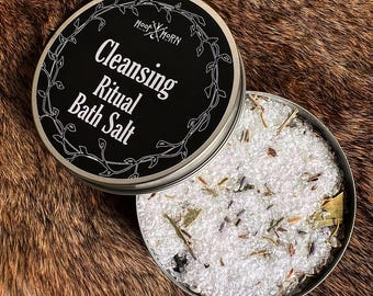 May include: A round, open tin of bath salts with the lid slightly ajar. The lid is black with white text that reads "Cleansing Ritual Bath Salt" and a decorative border. The bath salts are white with dried herbs and flowers.