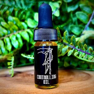 May include: A small glass dropper bottle filled with a golden liquid. The bottle features a black label with a white line drawing of a hand manipulating a marionette puppet and the text CONTROLLING OIL printed in white capital letters.