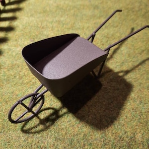 May include: A miniature metal wheelbarrow with a brown finish. The wheelbarrow is sitting on a green artificial grass surface.