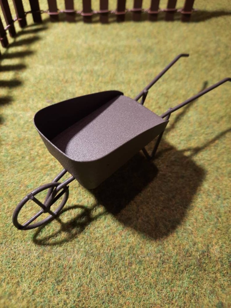 May include: A miniature metal wheelbarrow with a brown finish. The wheelbarrow is sitting on a green artificial grass surface.