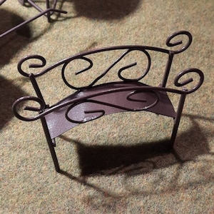 May include: A small, brown metal bench with a decorative, curved back and scrolled details. The bench is made of wrought iron and has a rustic, vintage look.