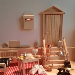 Könnte beinhalten: Ein Miniatur-Puppenhaus-Küchenset mit einer rot-weiß karierten Tischdecke, einem Holztisch mit einer Teekanne, einem Teller mit Essen und zwei Säcken Mehl mit der Aufschrift "Mehl 25 KGS". Es gibt einen hölzernen Briefkasten mit einem roten Umschlag darin, eine Holztür und einen Satz Kupfertöpfe auf einer Holztreppe.