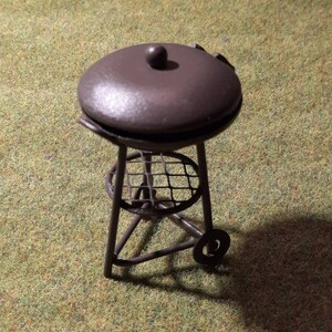 May include: A miniature black metal grill with a round lid and a wire rack. The grill has three legs and a wheel on one side.