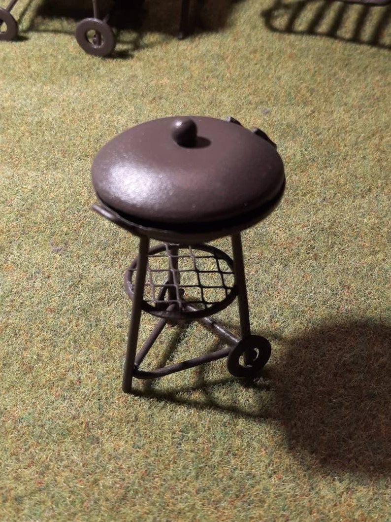 May include: A miniature black metal grill with a round lid and a wire rack. The grill has three legs and a wheel on one side.